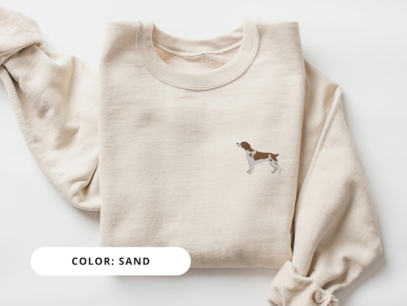 May include: A sand-colored sweatshirt with a brown and white dog illustration on the front. The text "COLOR: SAND" is printed below the illustration.