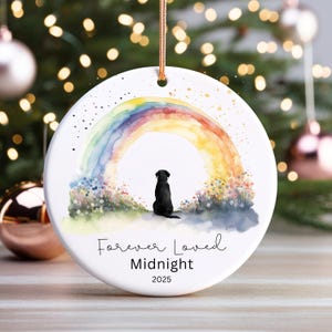 May include: A white ceramic ornament featuring a watercolor rainbow and a black dog silhouette. The text "Forever Loved Midnight 2025" is written below the image. A gold string is attached for hanging.