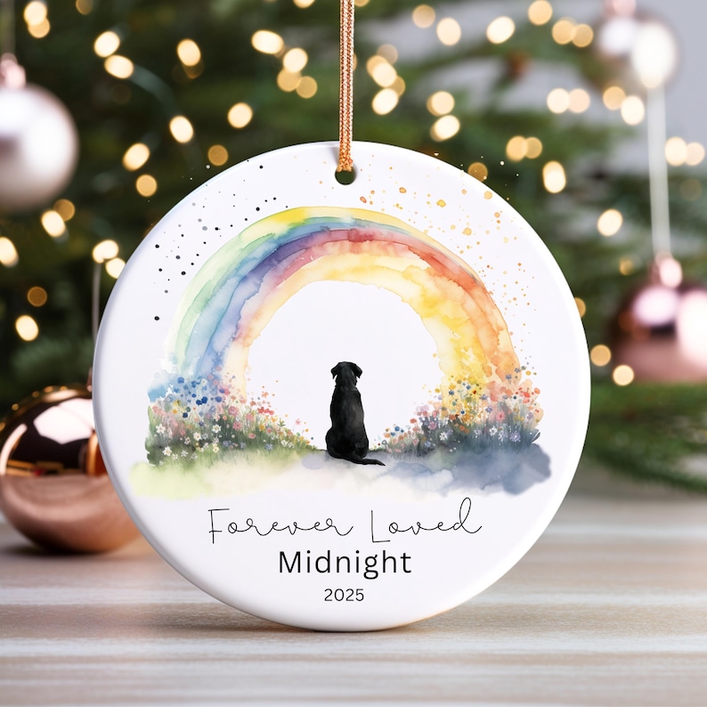 May include: A white ceramic ornament featuring a watercolor rainbow and a black dog silhouette. The text "Forever Loved Midnight 2025" is written below the image. A gold string is attached for hanging.