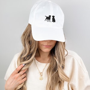 Op de afbeelding: Witte baseballpet met zwarte geborduurde silhouetten van drie honden. De pet heeft een gebogen klep en een verstelbare band.