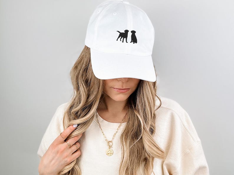 May include: White baseball cap with black embroidered silhouettes of three dogs. The cap has a curved brim and an adjustable strap.
