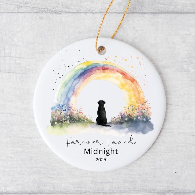 May include: A white ceramic ornament with a watercolor rainbow design and a black silhouette of a dog. The text "Forever Loved Midnight 2025" is written below the dog and rainbow. A gold string is attached for hanging.