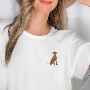 Pode incluir: Uma camiseta branca com uma silhueta de cachorro marrom na frente. O cachorro está sentado com a cabeça levantada.
