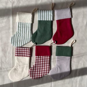 May include: Six Christmas stockings with hanging loops. Top left: cream with green stripes. Top middle: green with green striped cuff. Top right: red with plaid cuff. Bottom left: cream with red gingham cuff. Bottom middle: red with red gingham body. Bottom right: plaid with green cuff.