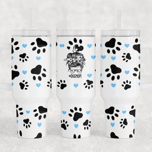 Op de afbeelding: Een witte roestvrijstalen beker met een patroon van zwarte poot afdrukken en blauwe harten. De beker heeft een deksel en een rietje. Op de beker staat "#DOGMOM".