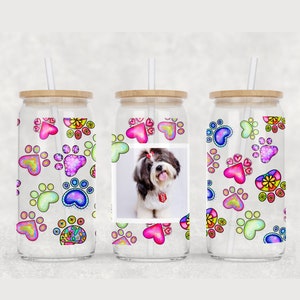 Puede incluir: Tres vasos de vidrio transparente con tapas de bambú y pajitas. Cada vaso presenta un patrón colorido de huellas de patas y corazones. El vaso del centro tiene una foto de un perro pequeño blanco con pelo negro en la cabeza y la cola.