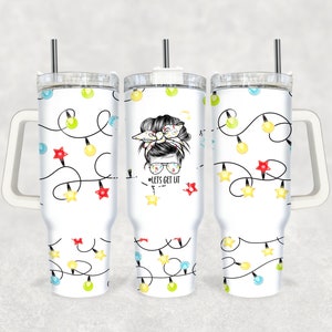 May include: A white tumbler with a handle and a straw. The tumbler has a design of colorful Christmas lights and a woman with a bun and glasses. The text "#LETS GET LIT" is on the tumbler.