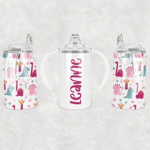 Puede incluir: Un vaso de acero inoxidable blanco con dos asas y una tapa de paja. El vaso está decorado con un patrón de dinosaurios rosa, azul y verde. El vaso está personalizado con el nombre "Leanne" en letras rosas.