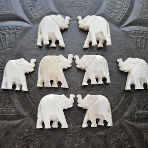 Puede incluir: Una colección de pequeñas figuras de elefantes talladas de color marfil dispuestas sobre una superficie oscura y texturizada. Los elefantes se enfrentan en parejas, mostrando sus trompas y detalles. El fondo tiene un patrón decorativo.