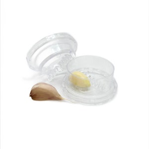 May include: Two clear plastic garlic presses with a single clove of garlic in one of the presses. A peeled garlic clove is also visible.
