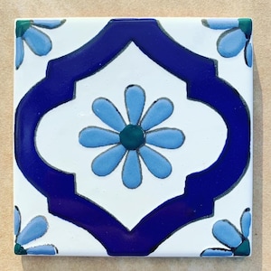 May include: A white ceramic tile with a blue and green floral design. The tile features a large blue flower with a green center and four smaller blue flowers in the corners. The tile is surrounded by a blue border.