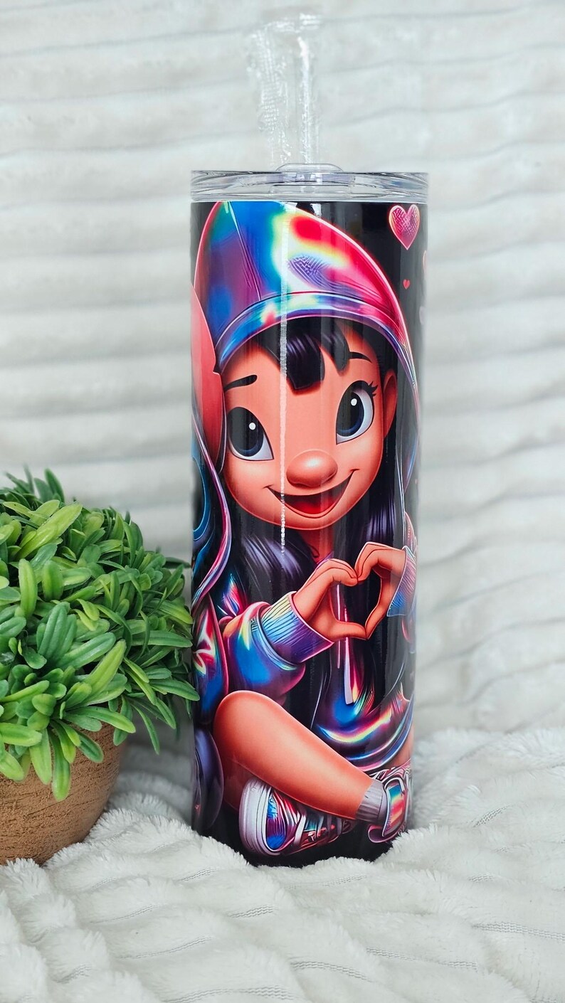 May include: A tall, cylindrical tumbler featuring a cartoon character in a colorful hoodie making a heart shape with their hands. The design includes vibrant rainbow hues and small pink hearts against a black background. A clear straw is visible.