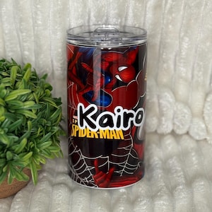 May include: A cylindrical tumbler with a Spider-Man design. The tumbler is mainly red with blue details, depicting the superhero. The name "Kairo" is in white, alongside the "Spider-Man" logo. The tumbler has a clear lid.