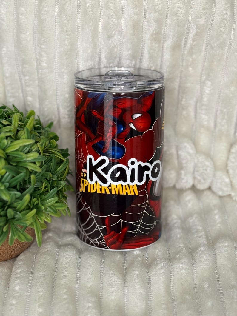 May include: A cylindrical tumbler with a Spider-Man design. The tumbler is mainly red with blue details, depicting the superhero. The name "Kairo" is in white, alongside the "Spider-Man" logo. The tumbler has a clear lid.
