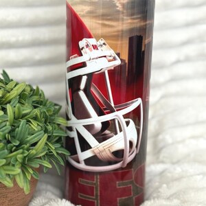 May include: A stainless steel tumbler with a red and white football helmet design. The background features a cityscape and sunset. The bottom of the tumbler has the letters "SF" in gold.