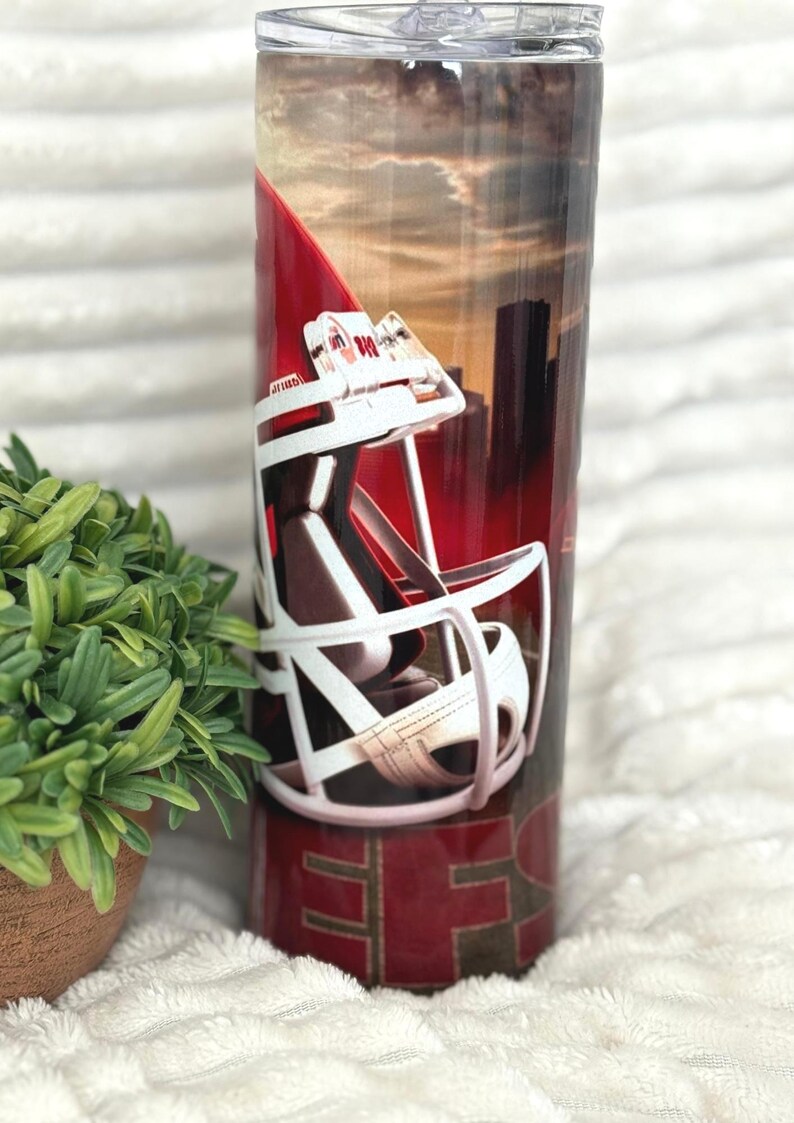 May include: A stainless steel tumbler with a red and white football helmet design. The background features a cityscape and sunset. The bottom of the tumbler has the letters "SF" in gold.