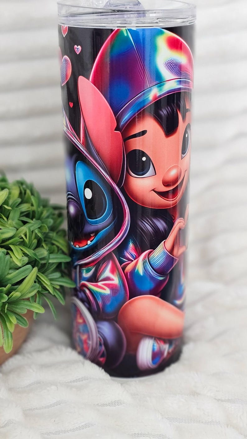 May include: A stainless steel tumbler featuring a vibrant illustration of Lilo and Stitch. The design showcases the characters in colorful, stylized outfits against a black background. The tumbler has a clear lid and is suitable for cold beverages.