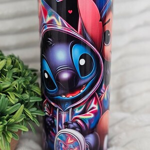 May include: A black tumbler featuring a colorful cartoon character design. The design includes a blue alien-like creature with large eyes and a hoodie, along with heart-shaped accents. The tumbler has a clear lid.