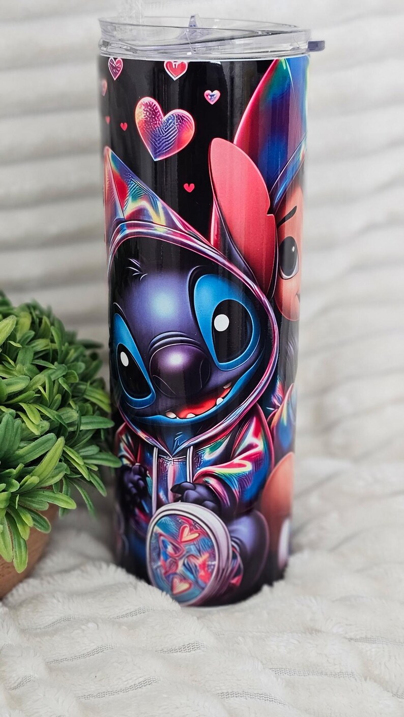 May include: A black tumbler featuring a colorful cartoon character design. The design includes a blue alien-like creature with large eyes and a hoodie, along with heart-shaped accents. The tumbler has a clear lid.
