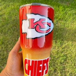 May include: A red, yellow, and white tumbler with the Kansas City Chiefs logo and the word "CHIEFS". The tumbler has a gradient design, transitioning from red to yellow. The Chiefs logo is white with red accents.