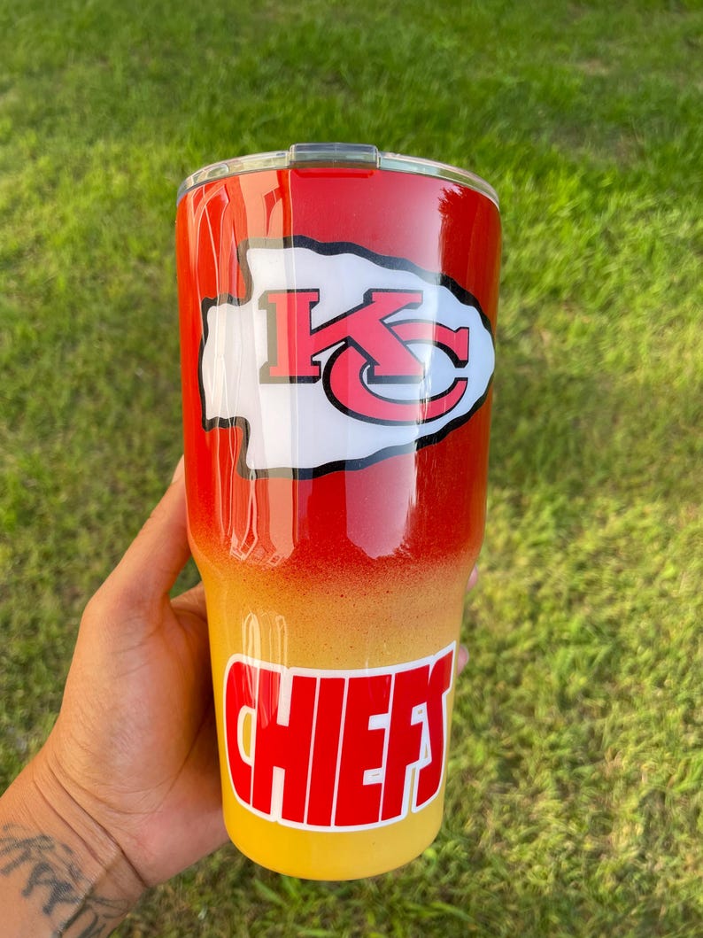 May include: A red, yellow, and white tumbler with the Kansas City Chiefs logo and the word "CHIEFS". The tumbler has a gradient design, transitioning from red to yellow. The Chiefs logo is white with red accents.