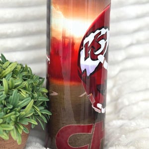 May include: A stainless steel tumbler with a red and white football helmet design, featuring the letters "KC" and the word "CHIEFS" in red. The background shows a sunset over a field. The tumbler is cylindrical and has a clear lid.