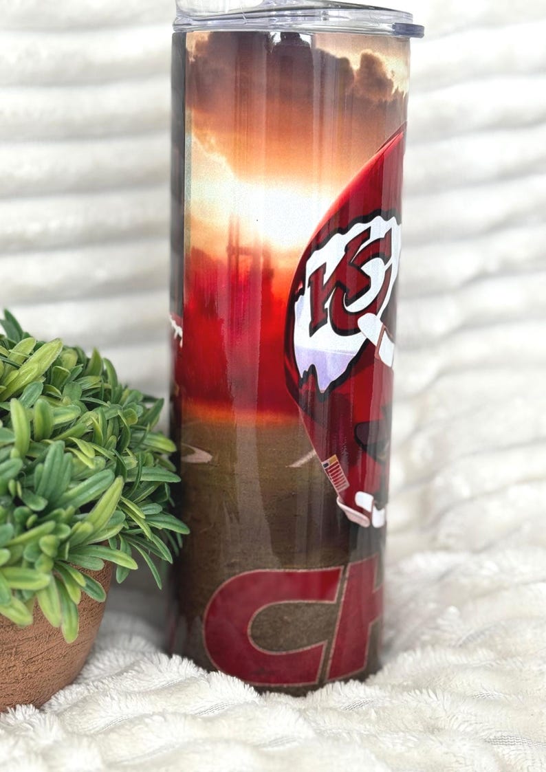 May include: A stainless steel tumbler with a red and white football helmet design, featuring the letters "KC" and the word "CHIEFS" in red. The background shows a sunset over a field. The tumbler is cylindrical and has a clear lid.