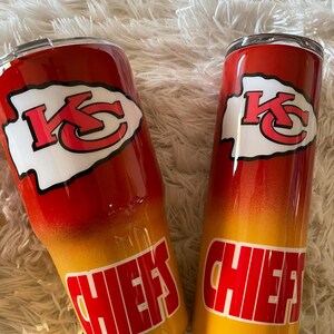 May include: Two stainless steel tumblers with a red, yellow, and orange gradient design. Each tumbler features the Kansas City Chiefs logo in white and red, with the word "CHIEFS" in red block letters.