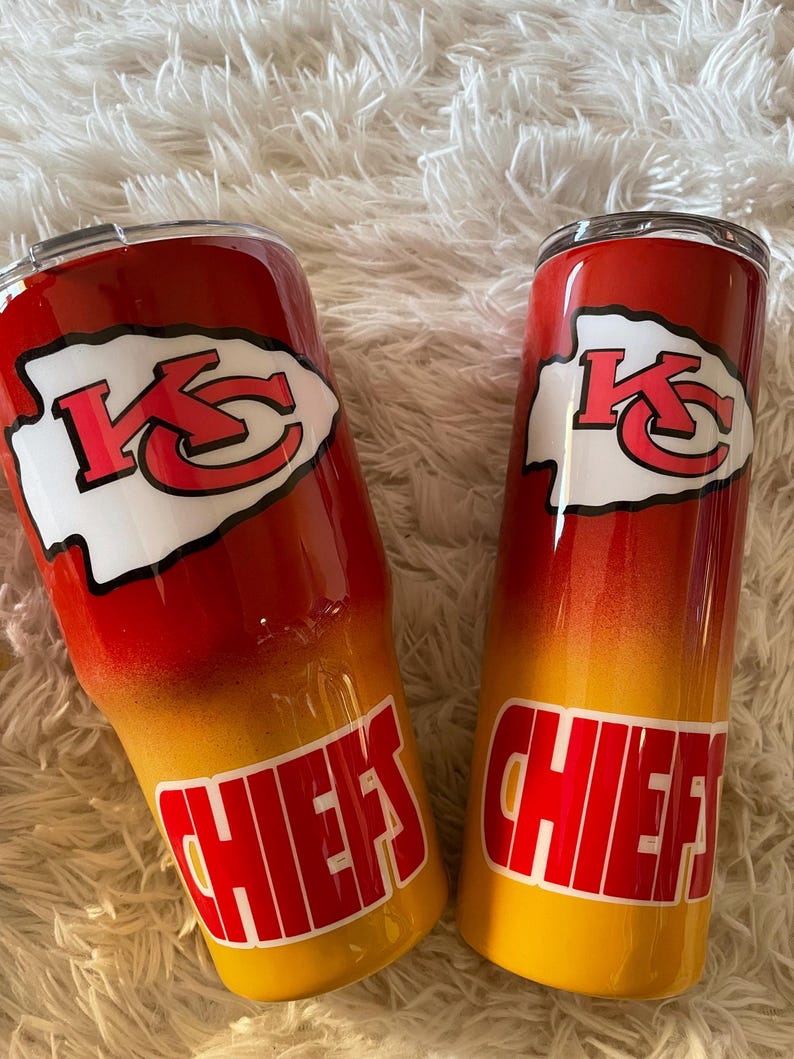 May include: Two stainless steel tumblers with a red, yellow, and orange gradient design. Each tumbler features the Kansas City Chiefs logo in white and red, with the word "CHIEFS" in red block letters.