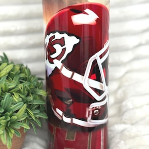 May include: A 590 ml insulated tumbler with a red and white football helmet design. The tumbler has a clear lid and a sunset background. The bottom of the tumbler features the letters "KC" in a red and gold pattern.