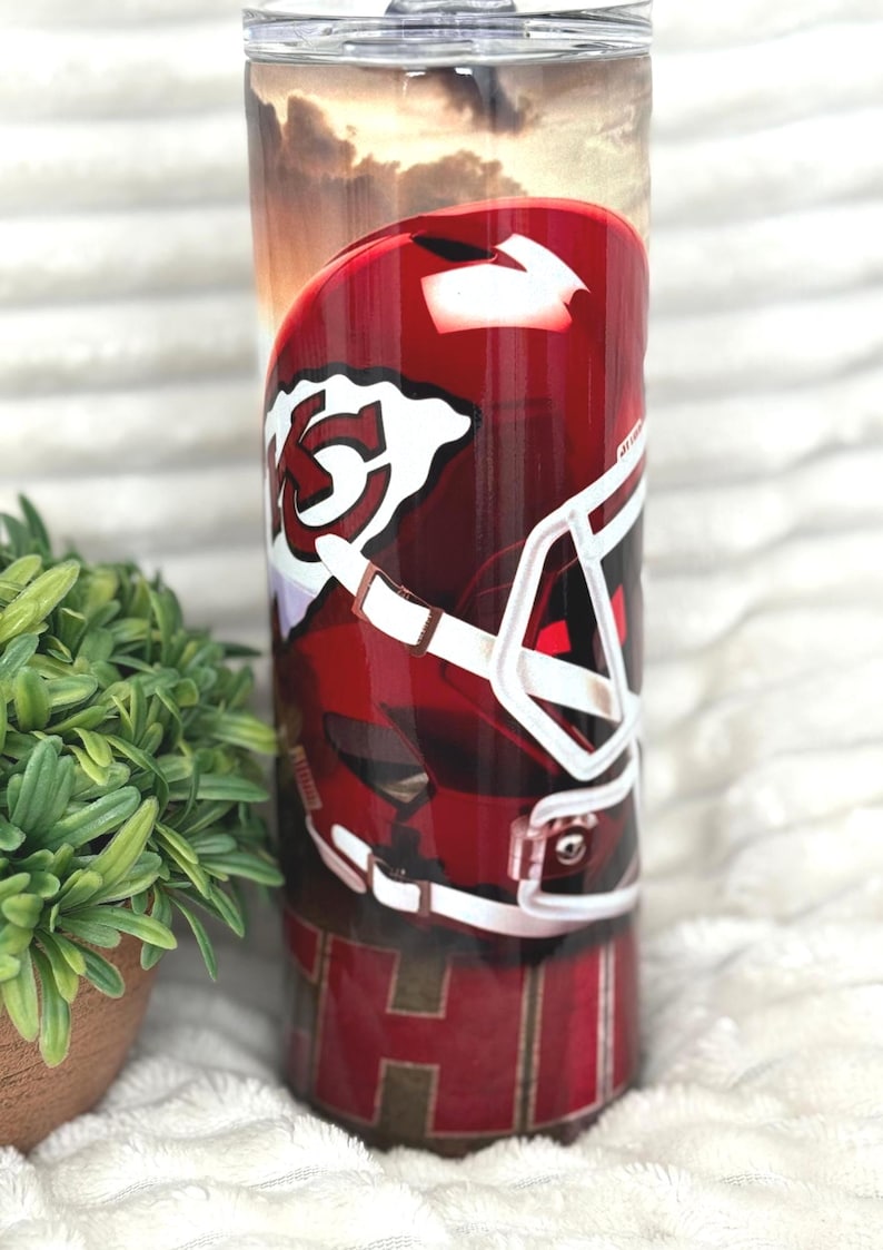 May include: A 590 ml insulated tumbler with a red and white football helmet design. The tumbler has a clear lid and a sunset background. The bottom of the tumbler features the letters "KC" in a red and gold pattern.