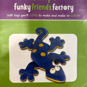 May include: A blue and yellow plush gecko toy kit from Funky Friends Factory. The kit includes a soft toy gecko with a heat pack, measuring 17 inches. The packaging features the text "Soft toys you'll LOVE to make and make to LOVE!" and "Gertie GECKO."