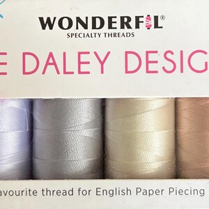 May include: A box of Wonderfil specialty threads, Sue Daley Designs, featuring six spools in various colors: black, lavender, silver, cream, peach, and brown. The text on the box reads "Sue Daley's favourite thread for English Paper Piecing and Appliqué."