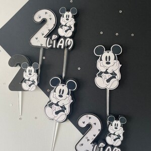 Puede incluir: Toppers para pastel en blanco y negro con Mickey Mouse y el n&uacute;mero dos. Los toppers est&aacute;n decorados con brillantina y el nombre "Liam" est&aacute; escrito en uno de los toppers.