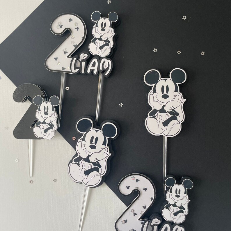 Puede incluir: Toppers para pastel en blanco y negro con Mickey Mouse y el n&uacute;mero dos. Los toppers est&aacute;n decorados con brillantina y el nombre "Liam" est&aacute; escrito en uno de los toppers.