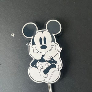 Puede incluir: Figura de dibujos animados de Mickey Mouse en blanco y negro con una cara sonriente. La figura est&aacute; sentada con las manos juntas frente a ella.