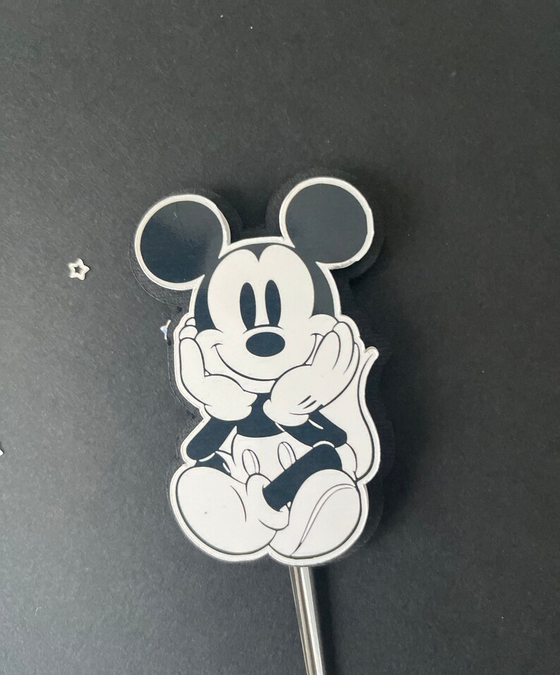 Puede incluir: Figura de dibujos animados de Mickey Mouse en blanco y negro con una cara sonriente. La figura est&aacute; sentada con las manos juntas frente a ella.