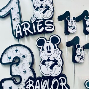 Puede incluir: Toppers para pastel en blanco y negro con Mickey Mouse, los n&uacute;meros 1 y 3, y los nombres "Aries" y "Baylor".
