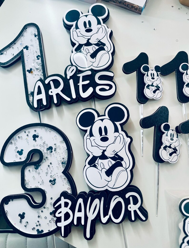 Puede incluir: Toppers para pastel en blanco y negro con Mickey Mouse, los n&uacute;meros 1 y 3, y los nombres "Aries" y "Baylor".