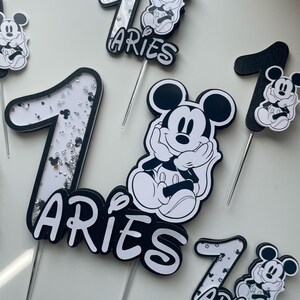 Puede incluir: Toppers para pastel en blanco y negro con Mickey Mouse y el n&uacute;mero uno. Los toppers tienen detalles de brillo y la palabra "Aries" escrita en ellos.