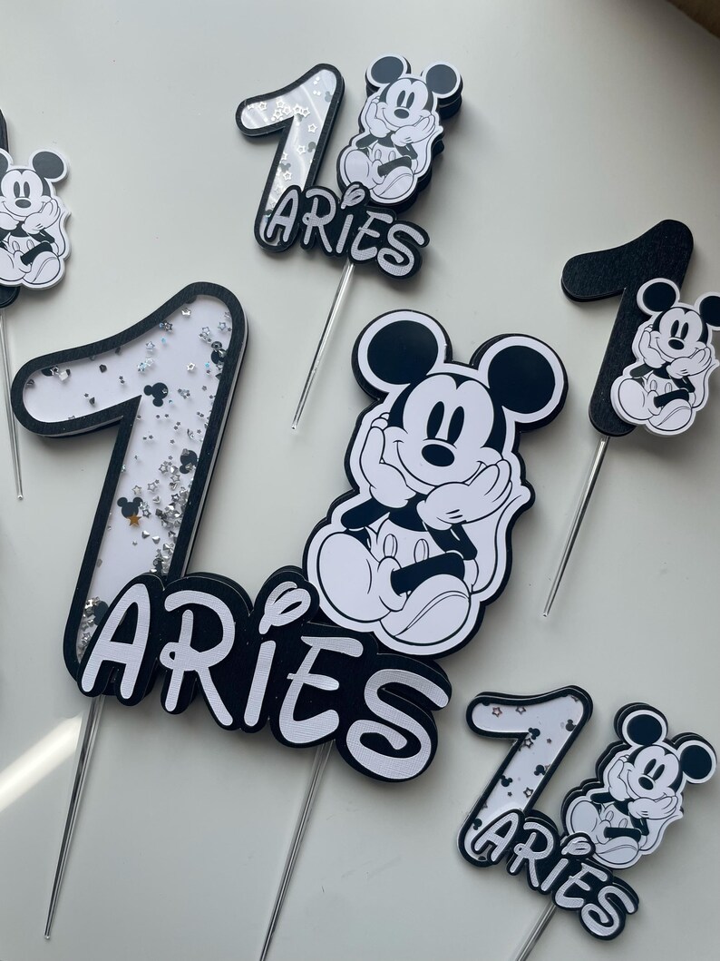 Puede incluir: Toppers para pastel en blanco y negro con Mickey Mouse y el n&uacute;mero uno. Los toppers tienen detalles de brillo y la palabra "Aries" escrita en ellos.