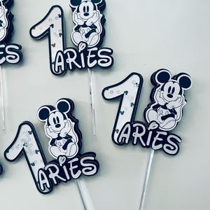 Puede incluir: Cuatro adornos para pasteles en blanco y negro con Mickey Mouse y el n&uacute;mero uno. Cada adorno tiene la palabra "Aries" escrita debajo del n&uacute;mero. Los adornos est&aacute;n en palos de pl&aacute;stico transparentes.