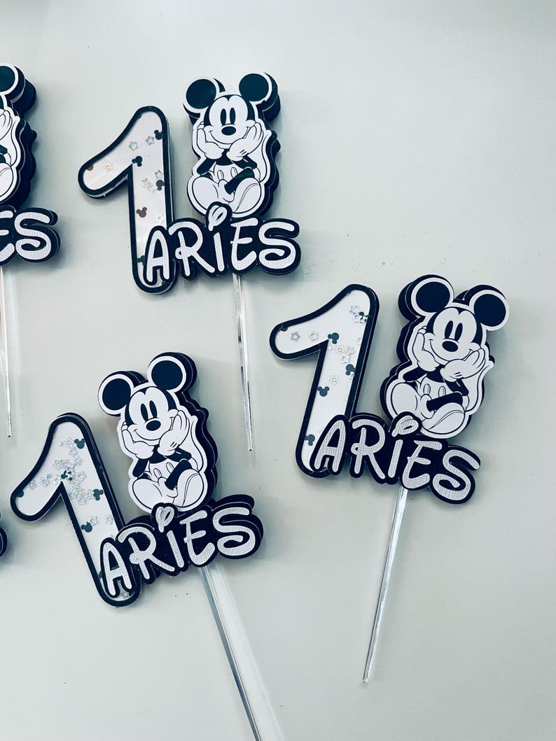 Puede incluir: Cuatro adornos para pasteles en blanco y negro con Mickey Mouse y el n&uacute;mero uno. Cada adorno tiene la palabra "Aries" escrita debajo del n&uacute;mero. Los adornos est&aacute;n en palos de pl&aacute;stico transparentes.