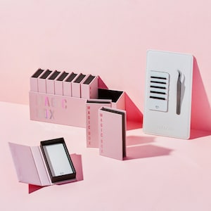 May include: A set of pink boxes with the text "MAGIC BOX" on the front. The boxes are arranged in a row and are stacked on top of each other. There is also a white tray with a set of false eyelashes and a pair of tweezers.