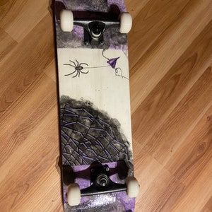 May include: A purple and black skateboard with a spider and flower design. The skateboard has black trucks and white wheels.