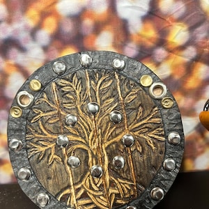 May include: A round, decorative shield with a tree of life design. The shield features a dark brown wooden base with a gold-colored tree design and silver and gold metal studs around the edge.