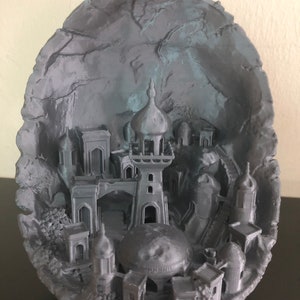 May include: A 3D printed model of a city inside a gray rock formation. The city features a tall tower with a domed roof and several smaller buildings.