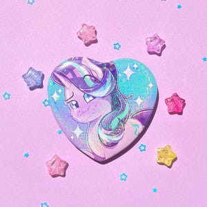 May include: A heart-shaped button featuring a cartoon pony with purple, blue, and teal hair. The button has a glittery finish and is surrounded by star-shaped beads on a lavender background. The pony is looking to the left.