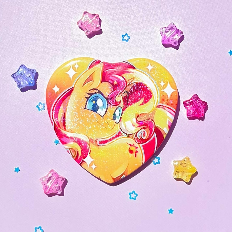 May include: A heart-shaped button featuring a cartoon pony with yellow and orange fur, pink and red hair, and blue eyes. The button is surrounded by pink, purple, and yellow star-shaped embellishments on a light purple background.