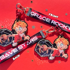May include: Two keychains with black fabric straps featuring a red and purple nebula pattern. One strap reads GRACE ROCKY, the other SAVE STARS. Each strap attaches to an acrylic charm showing a blonde character and a geometric shape with a creature.
